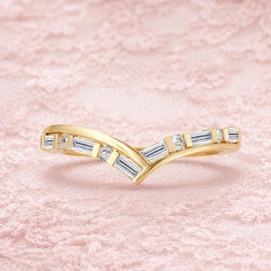 May include: A gold-colored ring with a V-shaped design. The ring is adorned with rectangular and round-cut clear gemstones. The ring is set against a soft pink, textured background.