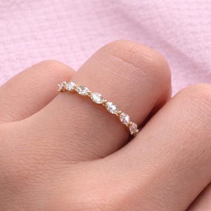 Puede incluir: Un anillo de oro con una serie de piedras preciosas claras de forma ovalada. El anillo está en un dedo, con un fondo rosa suave. Las piedras preciosas están espaciadas uniformemente y reflejan la luz.