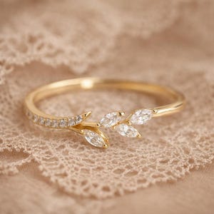 May include: A gold ring with a leaf motif, set with marquise-cut diamonds. The ring has a slender band and is displayed on a lace surface. The ring's design includes small, round diamonds along the band.