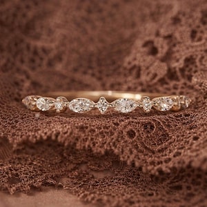 Puede incluir: Un delicado anillo de oro con una serie de gemas claras y brillantes. El diseño del anillo incorpora piedras de talla ovalada y redonda, dispuestas en un patrón repetitivo. El anillo se muestra sobre una superficie marrón texturizada, resaltando sus detalles.