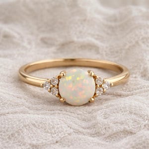 May include: A gold ring with a large, round opal centre stone. The opal is flanked by smaller, clear accent stones. The ring has a delicate band and is set against a soft, textured background.
