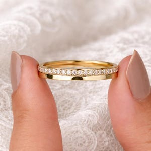 May include: Two gold rings, one with a band of small, clear gemstones. The rings are held between two fingers against a white, textured background. The rings are a bright, shiny gold color.
