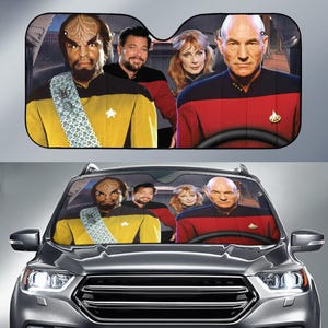 May include: Car sunshade featuring a group of people in Star Trek uniforms. The sunshade has a black background and a picture of the characters. The car sunshade is displayed in a gray car.