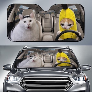 Op de afbeelding: Auto zonnescherm met een grappige afbeelding van twee katten in een auto. Eén kat draagt een bananenkostuum en rijdt, terwijl de andere er verbaasd uitziet. De achtergrond toont het interieur van de auto en een landschap.