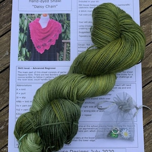 May include: A knitting pattern for a "Daisy Chain" shawl, featuring a hand-dyed green yarn with a pink shawl in the background. The pattern includes instructions for an advanced beginner knitter. The kit includes two skeins of yarn, stitch markers, and a project bag.