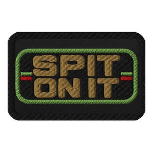 May include: A rectangular black embroidered patch featuring the text SPIT ON IT in tan thread. The text is enclosed within a bright green stitched border, with small red and green horizontal lines flanking the text on the left and right sides.
