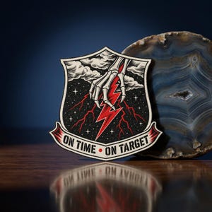 May include: A shield-shaped embroidered patch featuring a skeletal hand reaching down from clouds to grasp a bright red lightning bolt against a black starry sky. The bottom banner displays the text ON TIME • ON TARGET in bold black letters.