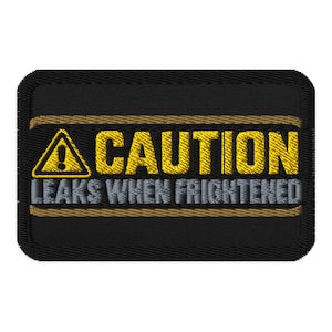 Op de afbeelding: Een rechthoekige zwarte geborduurde patch met een geel waarschuwingsdriehoek-icoon met een uitroepteken. De tekst CAUTION is in het geel geborduurd en de tekst LEAKS WHEN FRIGHTENED is in het grijs geborduurd, omrand door beige lijnen.