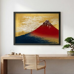 Op de afbeelding: Ingelijste kunstdruk met een gestileerde weergave van de berg Fuji. Het kunstwerk toont een felrode bergtop tegen een gouden achtergrond met witte wolkenformaties. Een donkerblauwe bosbasis maakt het landschap compleet.