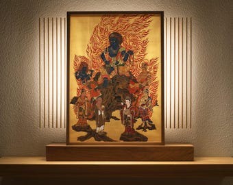 May include: A framed artwork depicting a deity surrounded by figures, set against a gold background with red flames. The artwork is displayed on a wooden base, with vertical light panels on either side, creating a warm, inviting ambiance. The artwork is a religious or spiritual piece.