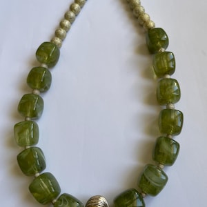 Peut inclure: Un collier composé de perles cubiques vert olive, semi-translucides, avec des motifs tourbillonnants blancs, alternées avec de petites perles d'espacement dorées texturées et une perle centrale sphérique dorée, nervurée et métallique.
