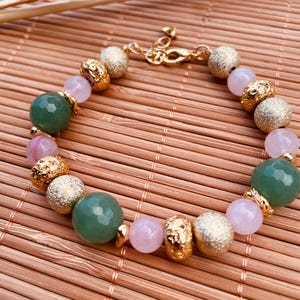 Peut inclure: Un bracelet de perles composé de perles vertes facettées, de perles rose clair lisses, de sphères métalliques dorées texturées et de pépites dorées irrégulières. Le bracelet est doté d'un fermoir mousqueton doré et d'une chaînette d'extension.