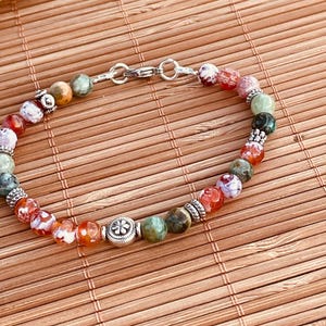 Puede incluir: Una pulsera de cuentas con piedras de ágata multicolor en tonos rojos, verdes y blancos, intercaladas con espaciadores de metal plateado y un dije central circular con diseño de flor, asegurada con un broche de langosta plateado.