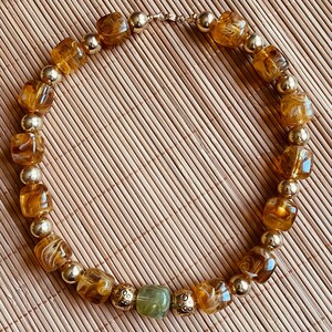 Peut inclure: Un collier de perles alternant des perles carrées marbrées couleur ambre et des perles sphériques en métal doré lisse. Le design comprend une perle carrée vert sauge au centre, entourée de deux perles dorées texturées, avec un fermoir doré.