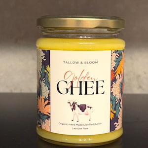 May include: A glass jar of Golden Ghee with a gold lid. The label features the text "Tallow & Bloom" and "Golden Ghee" with a cow illustration. The label also states "Organic Hand Made Clarified Butter" and "Lactose Free". The ghee is a bright yellow colour.
