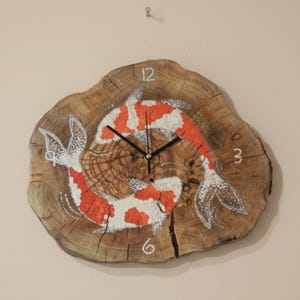 May include: A wooden wall clock with a natural wood grain finish. The clock face features two koi fish painted in red and white, with black clock hands and white numerals. The clock is round and measures approximately 12 inches in diameter.