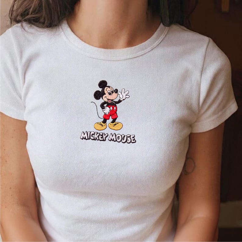 Op de afbeelding: Wit T-shirt met een geborduurd Mickey Mouse-ontwerp. De tekenfilmmuis wordt afgebeeld terwijl hij zwaait, met een rode broek, gele schoenen en zwarte handschoenen. De woorden "MICKEY MOUSE" zijn eronder in het zwart geborduurd.