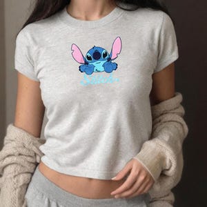 May include: A light grey t-shirt with a blue cartoon character and the word "Stitch" in light blue script. The character has large ears, a light blue body and pink ears. The text is in a cursive font.