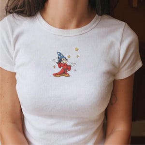 Könnte beinhalten: Weißes T-Shirt mit einem gestickten Bild von Mickey Mouse als Zauberer. Mickey trägt einen blauen Hut mit einem Halbmond, eine rote Robe und ist von gelben Sternen umgeben.