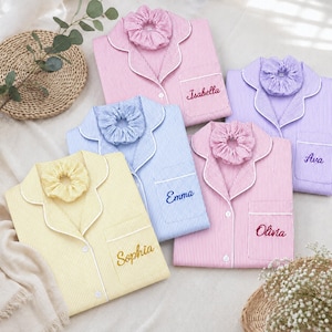 May include: Five folded button-down pajama shirts with matching scrunchies in pink, purple, blue, and yellow stripes. Each shirt features white piping, a collar, and a chest pocket embroidered with names: Isabella, Ava, Emma, Olivia, and Sophia.