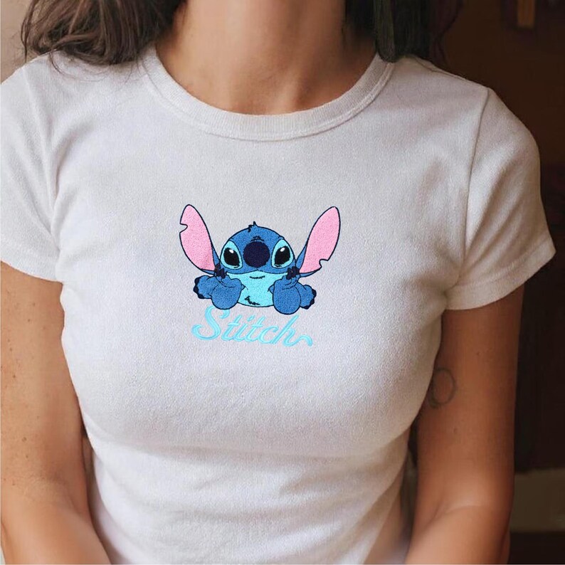 May include: White t-shirt with a blue cartoon character, large pink ears, and the word "Stitch" in light blue script. The character has a friendly expression. The t-shirt is a crew neck style.