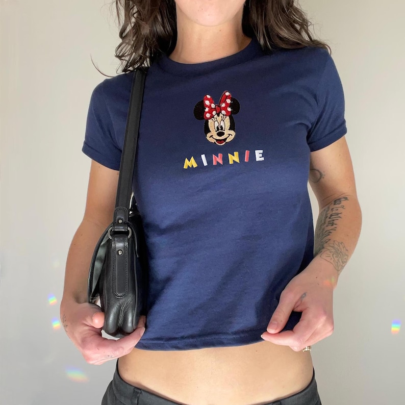 Pu&ograve; includere: T-shirt blu navy con un design ricamato di Minnie Mouse con un fiocco rosso e la scritta "MINNIE" a lettere colorate. La maglietta ha maniche corte e girocollo.