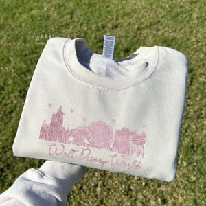 K&ouml;nnte beinhalten: Cremefarbenes Sweatshirt mit einem rosa gestickten Design der Skyline von Walt Disney World, einschlie&szlig;lich Cinderella's Castle und Spaceship Earth. Der Text "Walt Disney World" ist darunter gestickt.