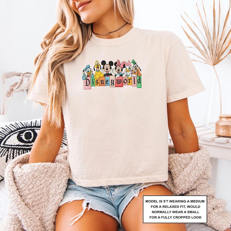 May include: A cream-coloured t-shirt with a colourful graphic of Disney characters and the text "Disneyworld". The graphic includes Mickey Mouse, Minnie Mouse, Donald Duck, and Goofy. The shirt has short sleeves and a relaxed fit.