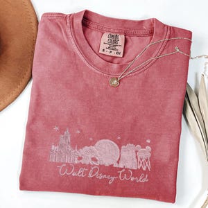 K&ouml;nnte beinhalten: Ein staubrosa Comfort Colors T-Shirt mit einer wei&szlig;en Grafik der Skyline von Walt Disney World. Das Shirt hat einen Rundhalsausschnitt und kurze &Auml;rmel. Eine silberne Halskette mit einem kleinen Anh&auml;nger liegt auf dem Shirt. Das Shirt ist gefaltet.