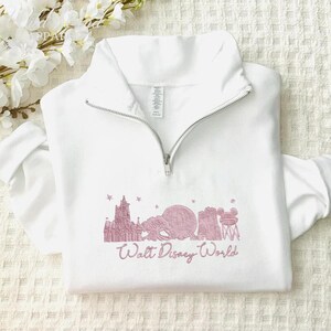 May include: White quarter-zip sweatshirt with a pink Walt Disney World design. The design features iconic park imagery and the words "Walt Disney World" in a script font. The sweatshirt has a silver zipper.