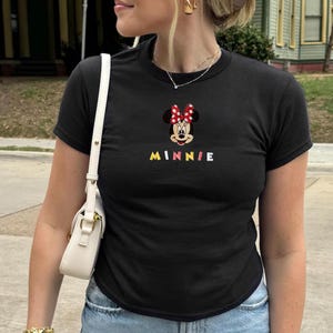 Pu&ograve; includere: T-shirt nera con un ricamo di Minnie Mouse, con un fiocco rosso a pois bianchi. La parola "MINNIE" &egrave; ricamata in lettere colorate sotto. La maglietta ha maniche corte e girocollo.
