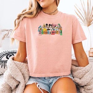 May include: A pale pink t-shirt with a graphic of Disney characters and the text "Disneyworld". The characters include Mickey Mouse, Minnie Mouse, Donald Duck, Goofy, and Daisy Duck. The shirt has short sleeves and a crew neck.