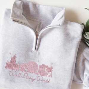 May include: Light gray quarter-zip sweatshirt with a pink embroidered Walt Disney World skyline design. The design includes Cinderella's castle, Spaceship Earth, and the iconic Mickey Mouse water tower. The sweatshirt has a high collar and a silver zipper.