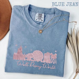 K&ouml;nnte beinhalten: Blaues Comfort Colors T-Shirt mit rosa gesticktem Walt Disney World-Design. Das Design umfasst Cinderella's Schloss, Spaceship Earth und den Tower of Terror. Eine goldene Halskette mit Anh&auml;nger liegt auf dem Shirt.