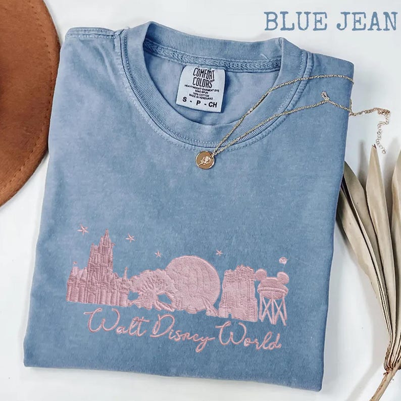 K&ouml;nnte beinhalten: Blaues Comfort Colors T-Shirt mit rosa gesticktem Walt Disney World-Design. Das Design umfasst Cinderella's Schloss, Spaceship Earth und den Tower of Terror. Eine goldene Halskette mit Anh&auml;nger liegt auf dem Shirt.