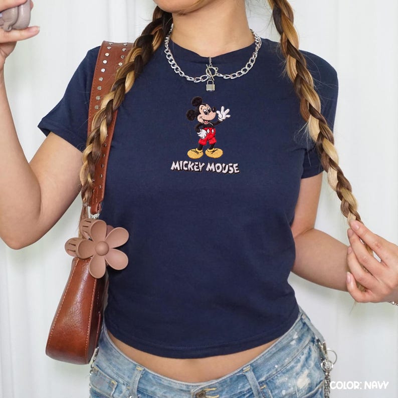 Op de afbeelding: Marineblauw T-shirt met een geborduurd Mickey Mouse-ontwerp. Het stripfiguur is afgebeeld in een rode broek, gele schoenen en witte handschoenen, met de woorden "Mickey Mouse" eronder geborduurd. Het shirt heeft een getailleerde stijl.
