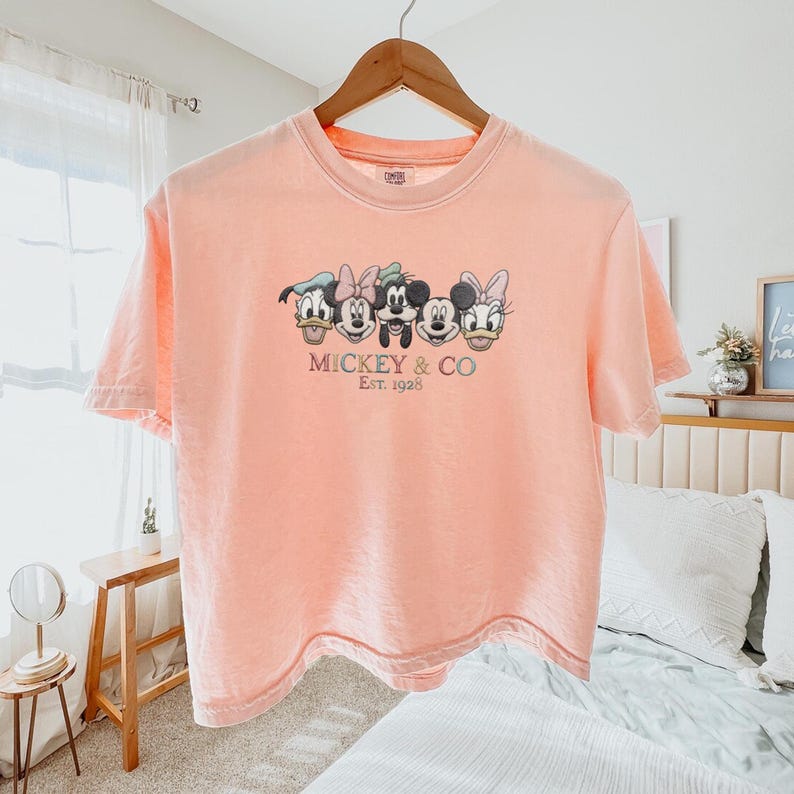 Op de afbeelding: Een perzikkleurig T-shirt met de gezichten van Mickey Mouse, Minnie Mouse, Donald Duck en Daisy Duck. De tekst "Mickey & Co Est. 1928" is onder de karakters geborduurd. Het shirt hangt aan een houten hanger.