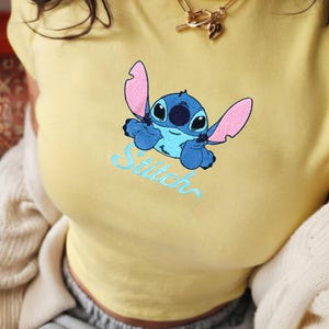 May include: A yellow t-shirt with an embroidered image of the character Stitch from the Disney film Lilo & Stitch. The blue Stitch is in a playful pose with the word "Stitch" embroidered in light blue script below.