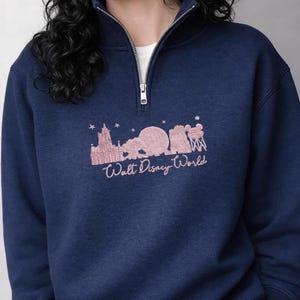 May include: Navy blue sweatshirt with a quarter-zip collar. The sweatshirt features a pink graphic of the Walt Disney World skyline and the words "Walt Disney World" in a cursive font. The sweatshirt has a white undershirt peeking out from the collar.