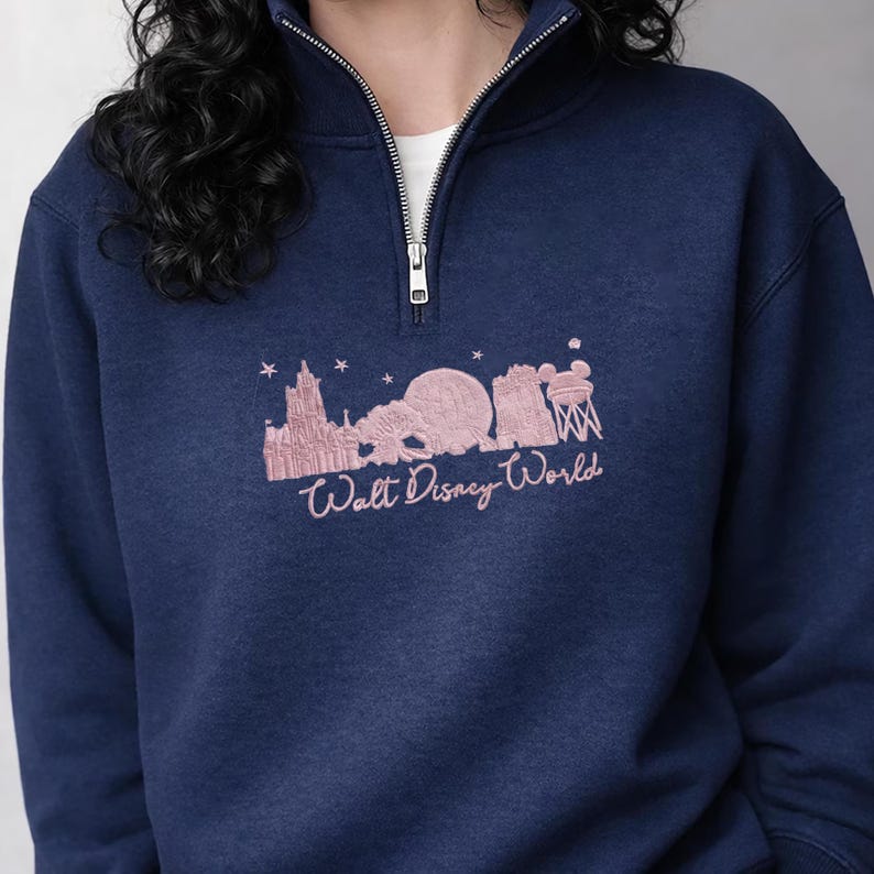 May include: Navy blue sweatshirt with a quarter-zip collar. The sweatshirt features a pink graphic of the Walt Disney World skyline and the words "Walt Disney World" in a cursive font. The sweatshirt has a white undershirt peeking out from the collar.
