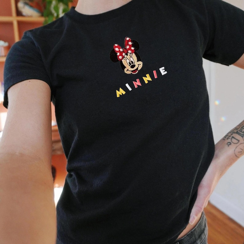 Pu&ograve; includere: T-shirt nera con un ricamo di Minnie Mouse, un fiocco rosso e il nome "MINNIE" in lettere colorate. Un design classico per i fan di Disney.