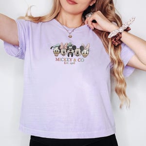 Op de afbeelding: Een lichtpaars t-shirt met een afbeelding van Mickey Mouse, Minnie Mouse, Donald Duck en Daisy Duck. De tekst "Mickey & Co. Est. 1928" staat eronder. Het shirt heeft korte mouwen.