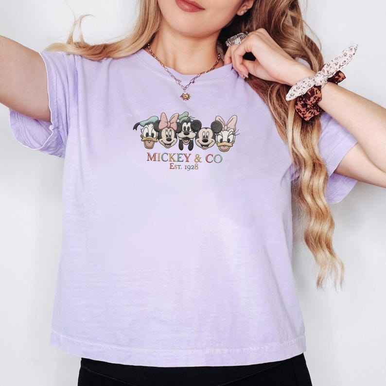 Op de afbeelding: Een lichtpaars t-shirt met een afbeelding van Mickey Mouse, Minnie Mouse, Donald Duck en Daisy Duck. De tekst "Mickey & Co. Est. 1928" staat eronder. Het shirt heeft korte mouwen.