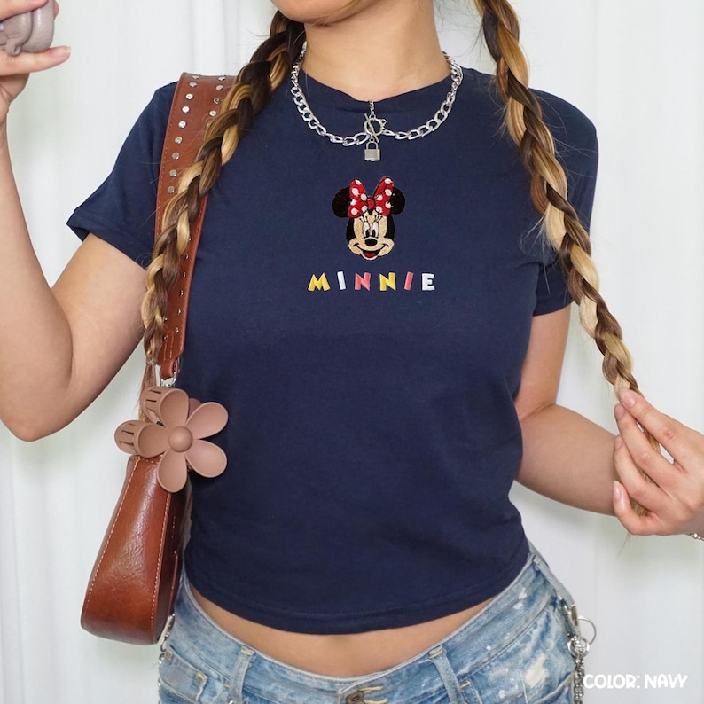 Pu&ograve; includere: T-shirt blu navy con un design ricamato di Minnie Mouse con fiocco rosso e il nome "MINNIE" a lettere colorate. Il top corto &egrave; abbinato a jeans blu. &Egrave; visibile anche una borsa marrone con un dettaglio floreale.