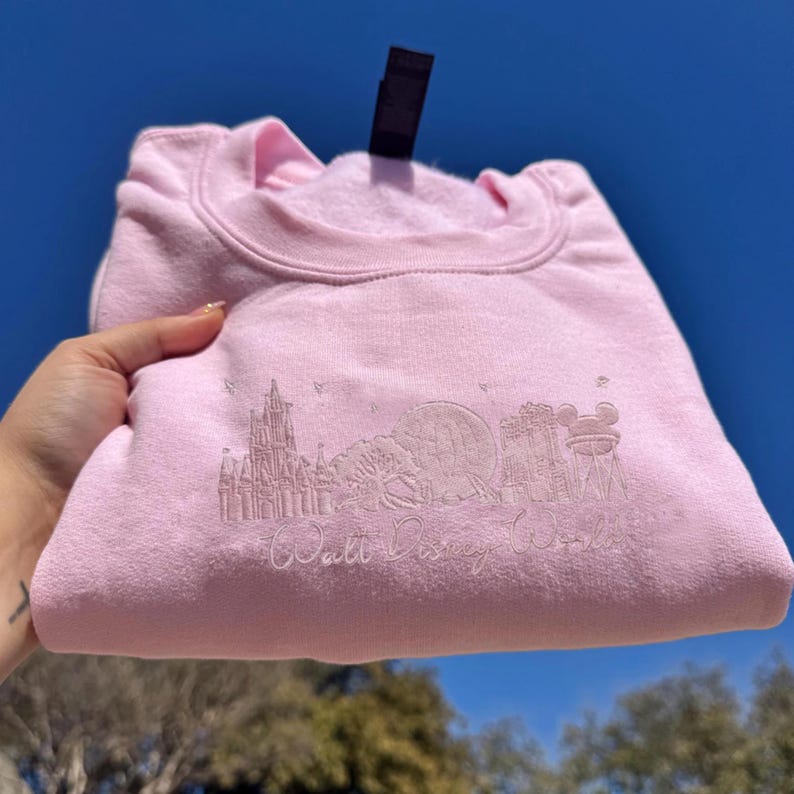 K&ouml;nnte beinhalten: Hellrosa Sweatshirt mit Rundhalsausschnitt. Auf der Vorderseite des Sweatshirts befindet sich ein gesticktes Design des Walt Disney World-Logos und verschiedener Parksymbole. Das Sweatshirt ist gefaltet und vor blauem Himmel gehalten.