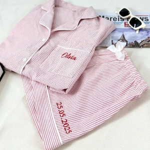 Könnte beinhalten: Rosa-weiß gestreifter Pyjama-Set mit weißer Paspelierung. Das Oberteil hat eine Brusttasche mit dem rot gestickten Namen "Olivia". Die Hose hat das Datum "25.05.2025" rot gestickt. Eine weiße Schleife und Sonnenbrille sind ebenfalls im Bild.