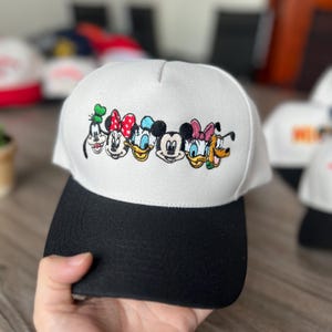 May include: A white and black baseball cap with embroidered cartoon character faces. The cap has a white crown and a black peak. The embroidered characters include Mickey Mouse, Minnie Mouse, Donald Duck, Daisy Duck, Goofy, and Pluto.
