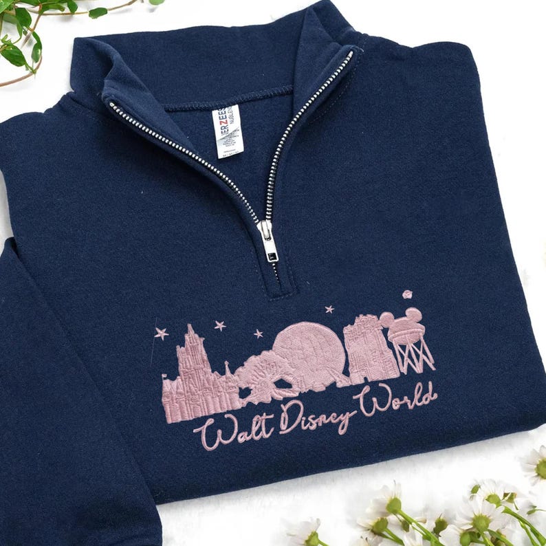 May include: Navy blue quarter-zip sweatshirt with a pink embroidered Walt Disney World design. The design includes the castle, Spaceship Earth, and other park icons. The sweatshirt has a silver zipper and a small tag.
