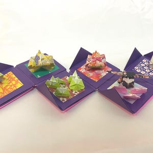 以下が含まれることがあります： さまざまな折り紙作品が入った、つながった紫色の折り紙の箱のシリーズ。作品には、折り紙の山、人物、その他の形があり、すべてさまざまな色と模様で表現されています。箱はジグザグ模様に配置されています。