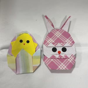 May include: Two origami figures: a yellow chick emerging from a pastel egg and a pink plaid bunny. The bunny has black eyes and a red nose. Both figures are made of folded paper.